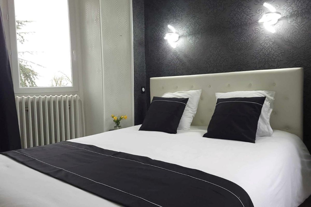 Une chambre d’hôtel avec un grand lit, du linge de lit blanc, deux oreillers noirs et blancs, un plaid noir et blanc, une tête de lit blanche et une fenêtre donnant sur des arbres.