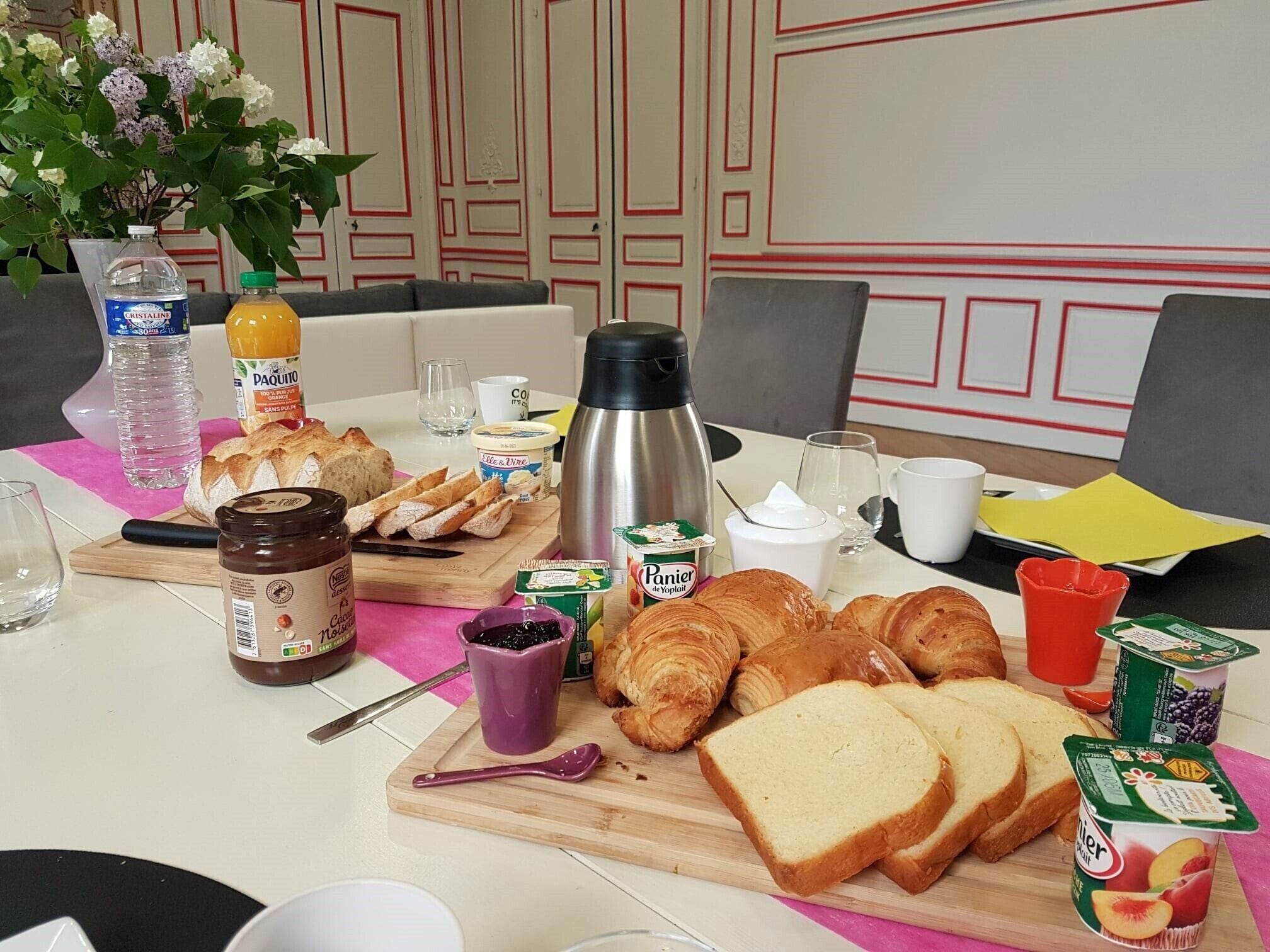 Une table de petit-déjeuner avec du pain, des viennoiseries, des condiments et des boissons.