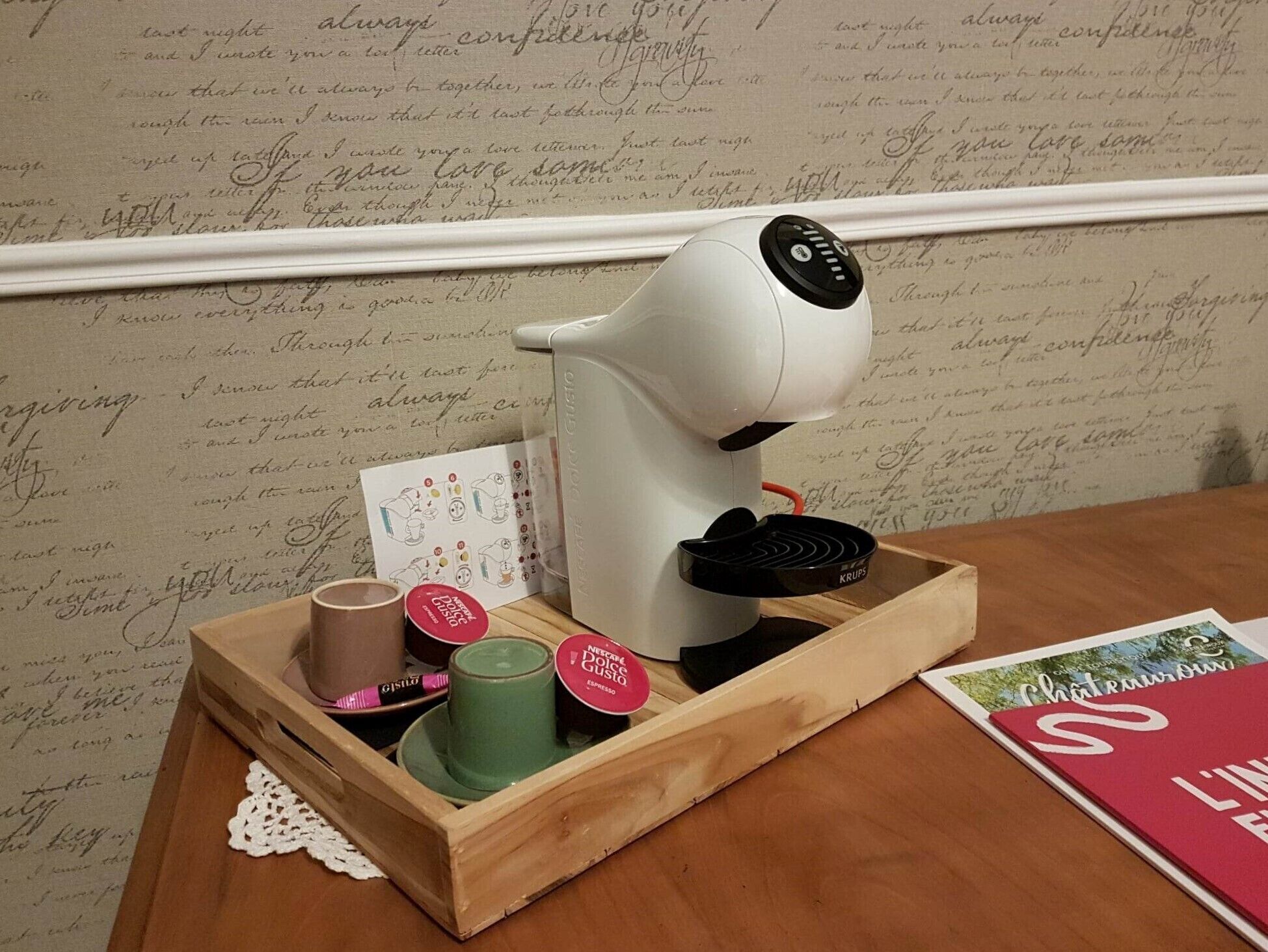 A coffee maker on a wooden tray with a doily, a green mug, and a pink mug.