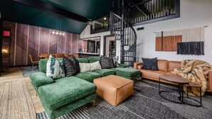 A modern living room with a green sectional sofa, a brown leather ottoman, and a tan sofa. A staircase with a black railing leads to an upper level. - Finger Lakes Escape • FLX Chalet + Private Pond (Branchport)