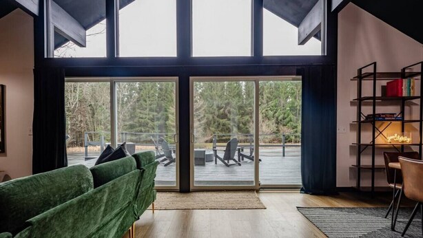 A modern living room with a green sofa, a wooden floor, and a large sliding glass door leading to an outdoor deck with chairs and a table. - Finger Lakes Escape • FLX Chalet + Private Pond (Branchport)