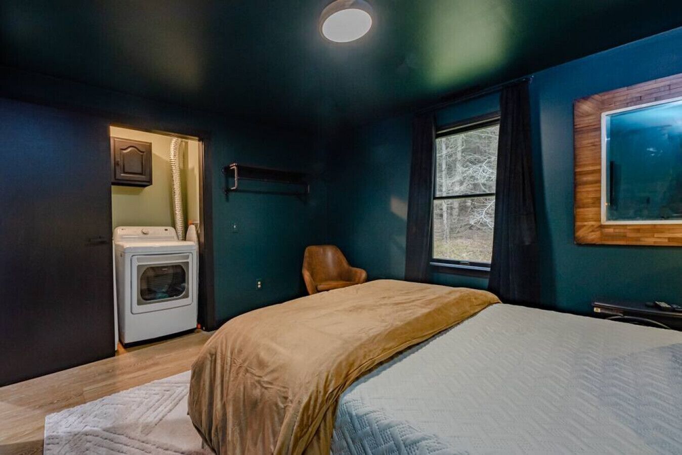 A bedroom with a bed, a chair, a washing machine, and a window with a view of trees.