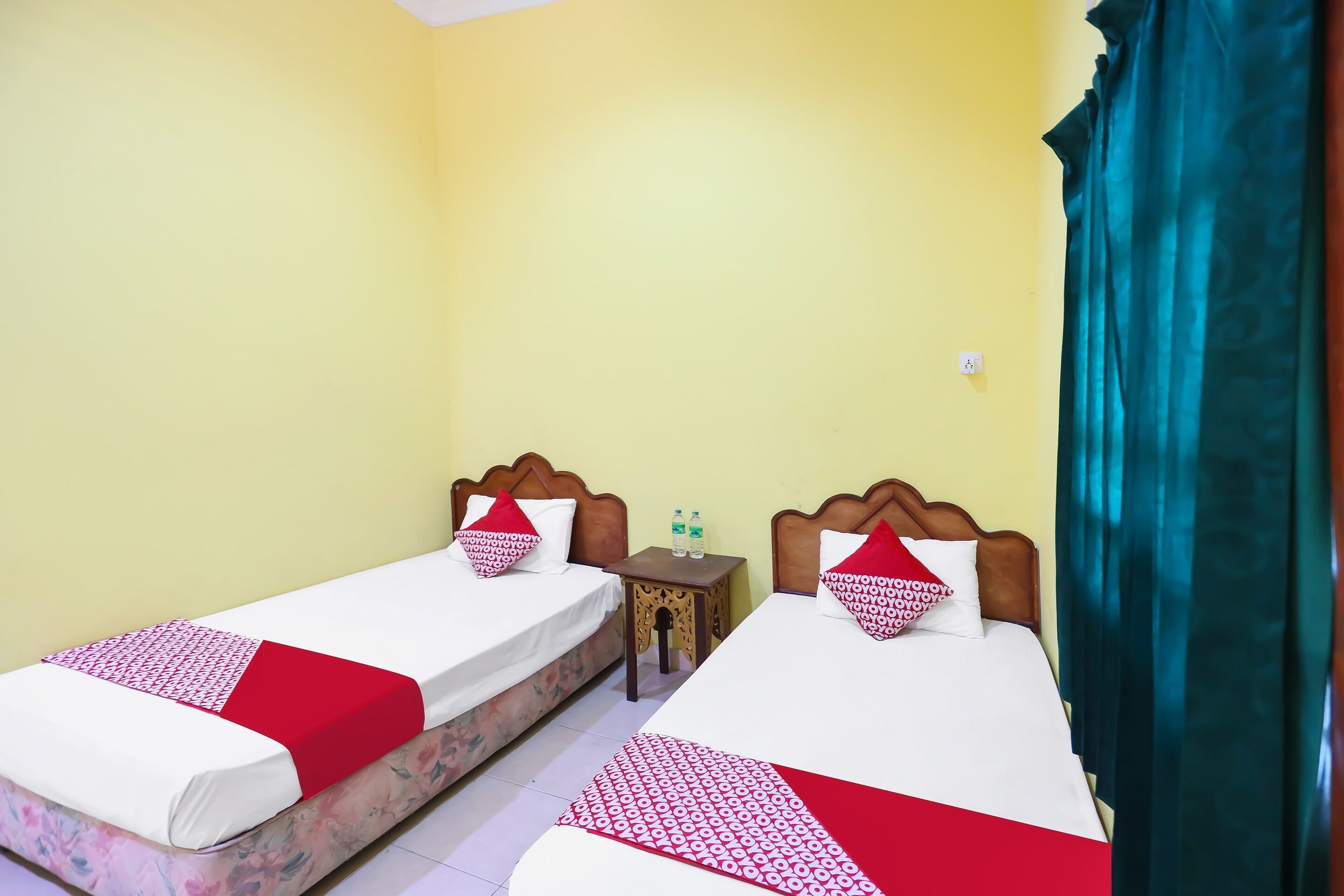 A hotel room with two single beds, each with a red and white patterned bedspread, wooden headboards, and matching pillows. There is a small wooden nightstand between the beds with two bottles of water. The room has yellow walls and a teal curtain on the right side.