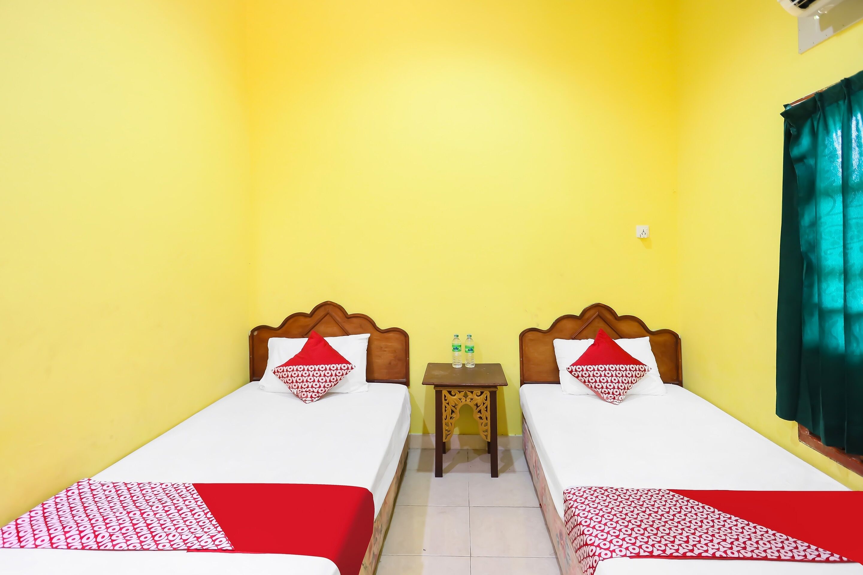 A room with two beds, each with a wooden headboard, white bedding, and red and white patterned bedspreads. There is a small table with two bottles of water between the beds.