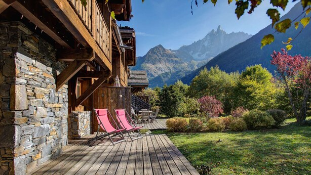 A wooden cabin with a stone wall, a wooden deck, and two red lounge chairs.