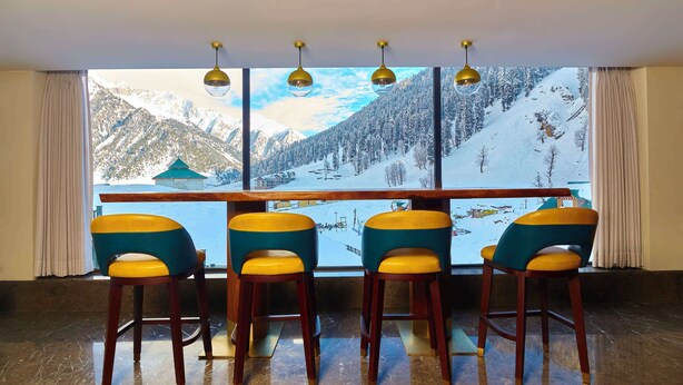 A room with a large window overlooking a snowy mountain landscape, wooden bar stools, and a ceiling light fixture. - Radisson Hotel Sonamarg (Gund)