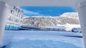 A snow-covered courtyard with a mountain backdrop, a building with large windows, and a clear blue sky. - Radisson Hotel Sonamarg (Gund)