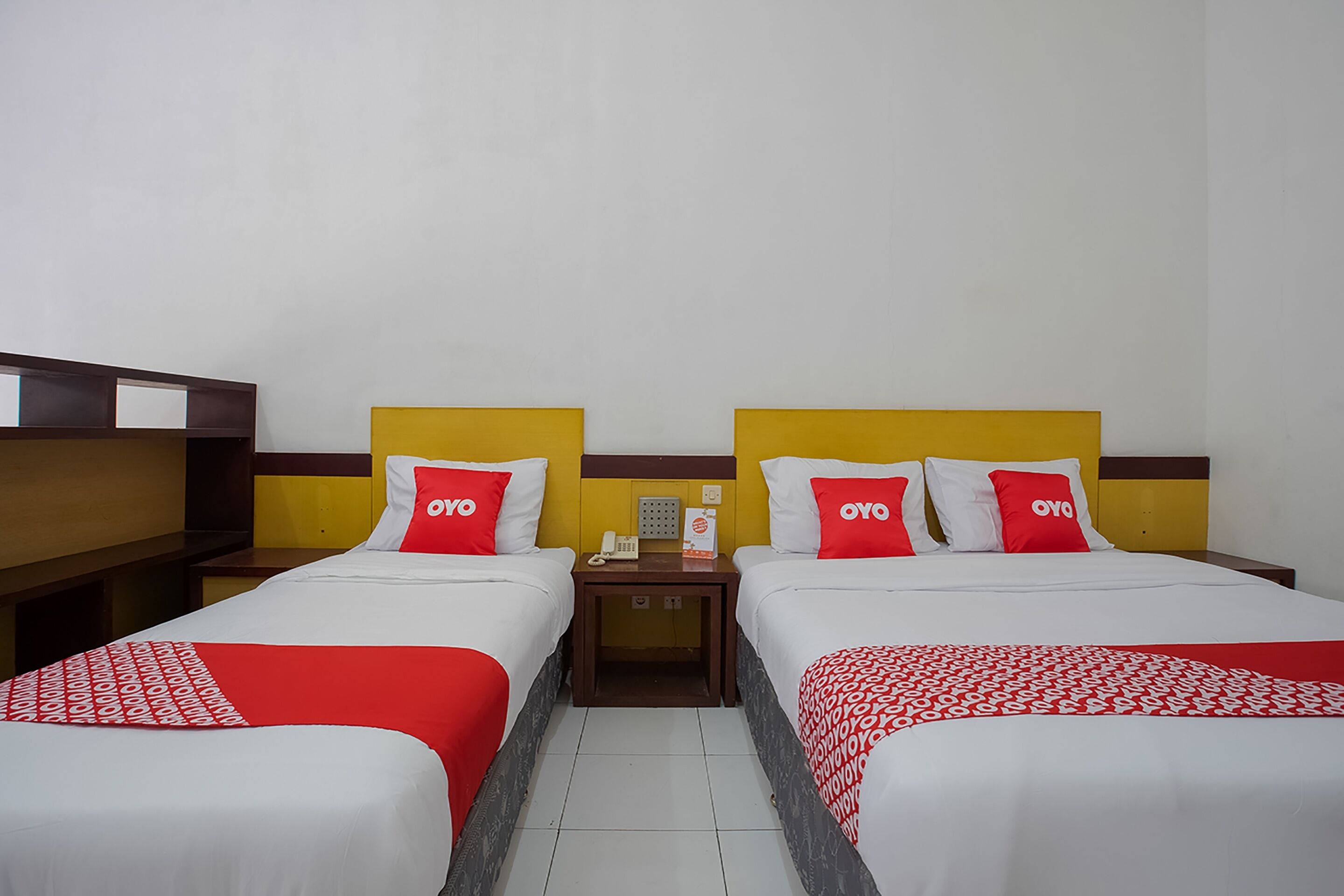 A hotel room with two beds, each with a red and white bedspread and matching pillows. There is a nightstand between the beds with a telephone and a small table.