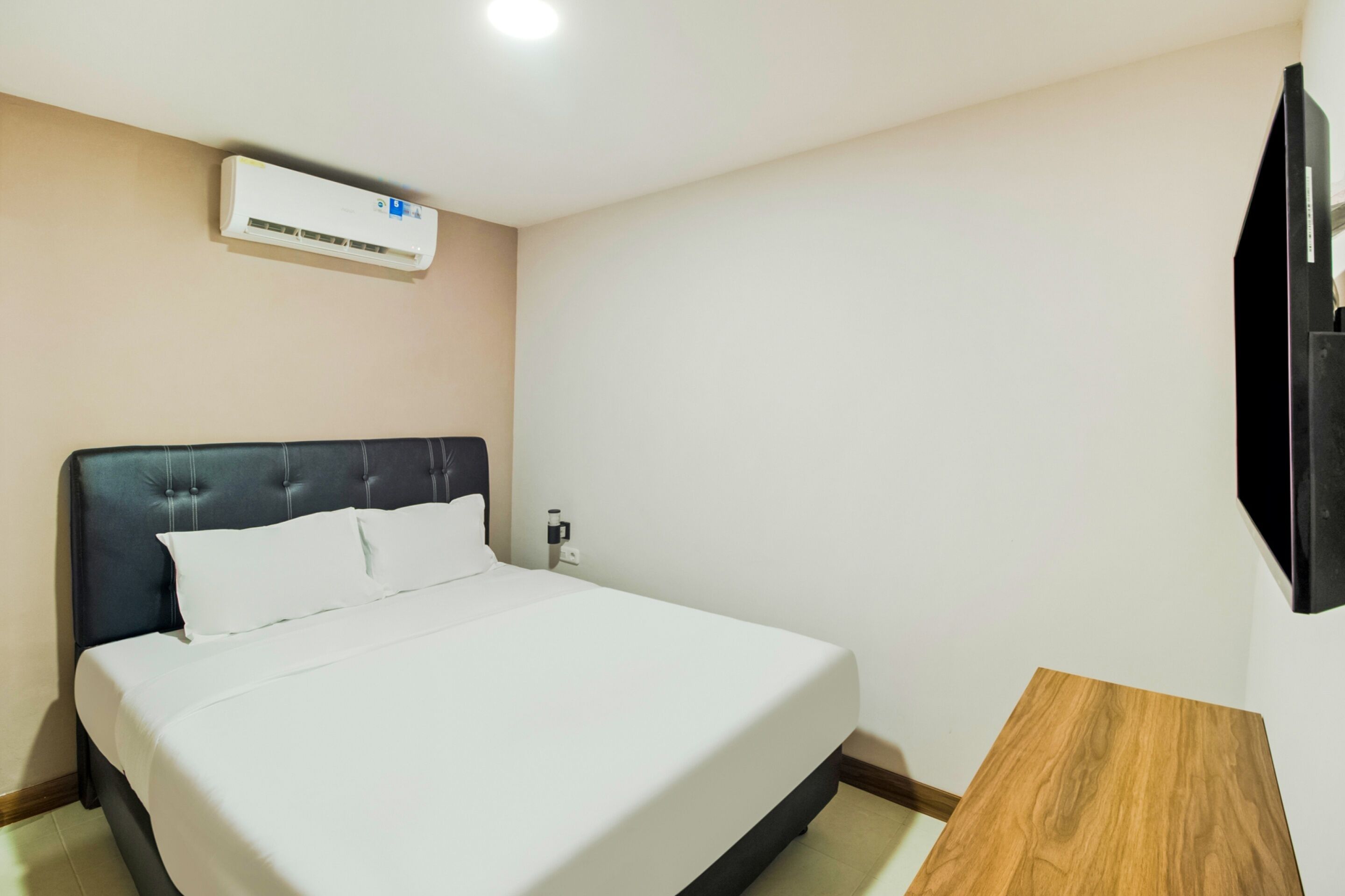 A hotel room with a bed, a wooden side table, a wall-mounted air conditioner, and a flat-screen TV.