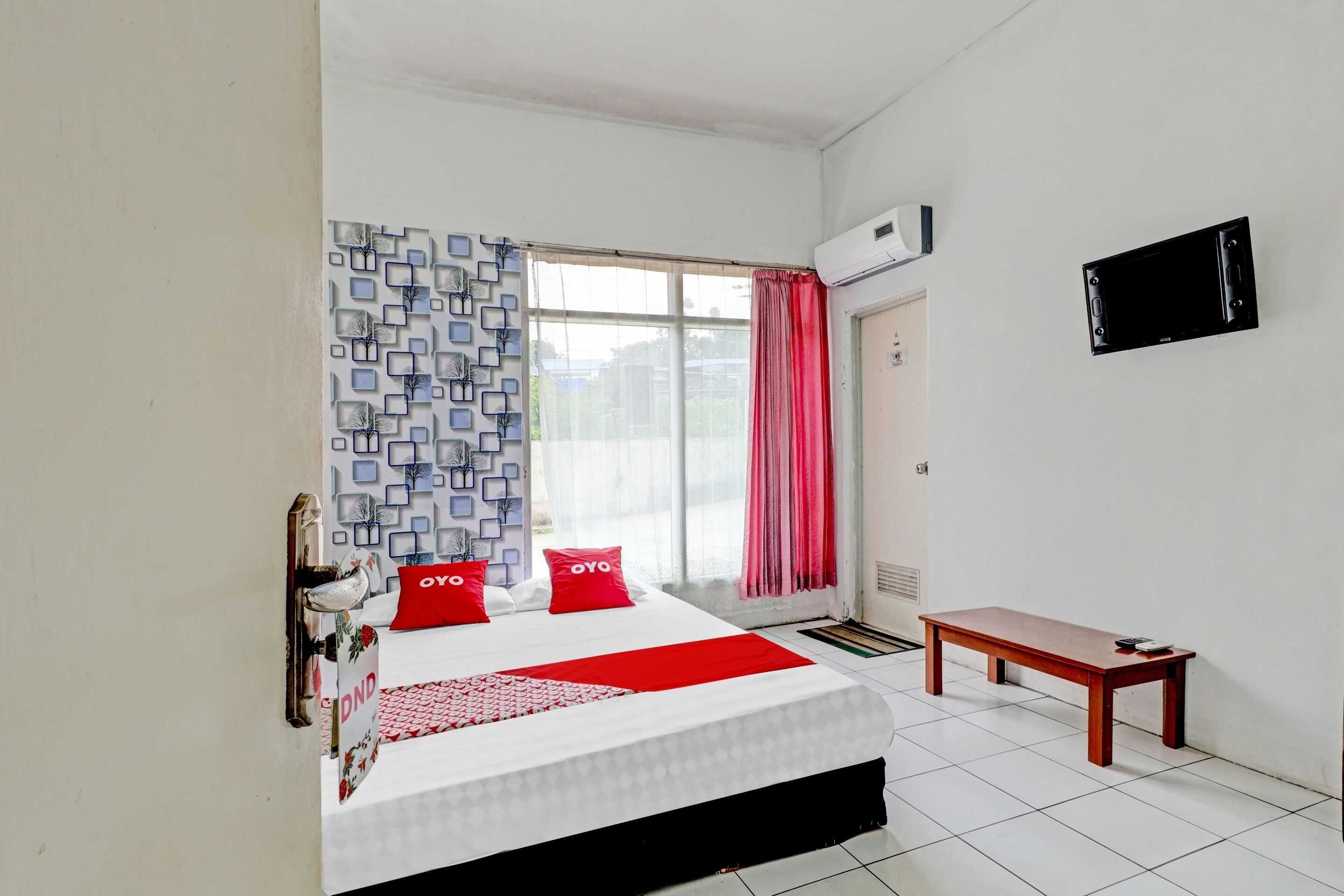 A hotel room with a bed, a bedside table, a window with curtains, and a wall-mounted TV.