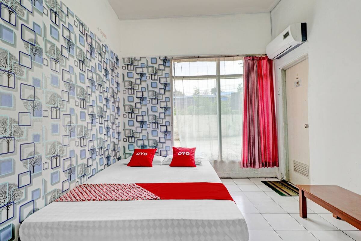 A hotel room with a bed, a bench, a window with curtains, and a wall with a geometric pattern.