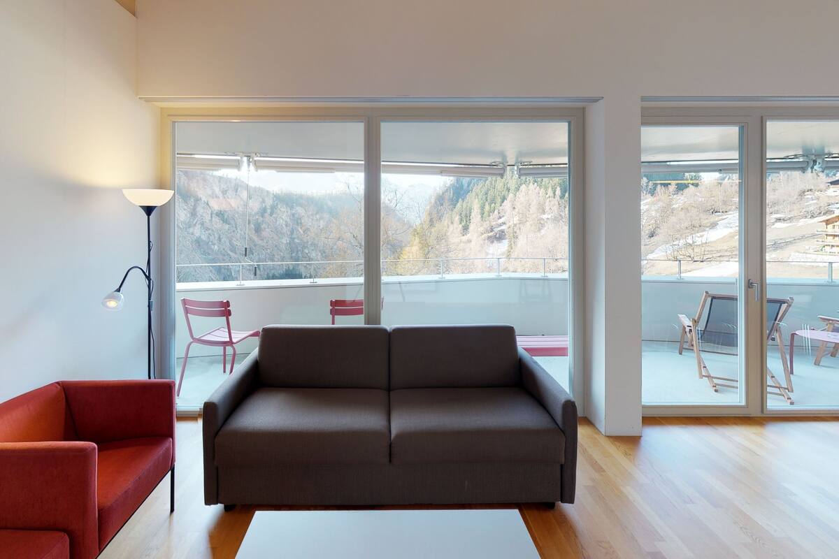 A modern living room with a sofa, a red chair, and a floor lamp. It features large windows with a view of a mountainous landscape.