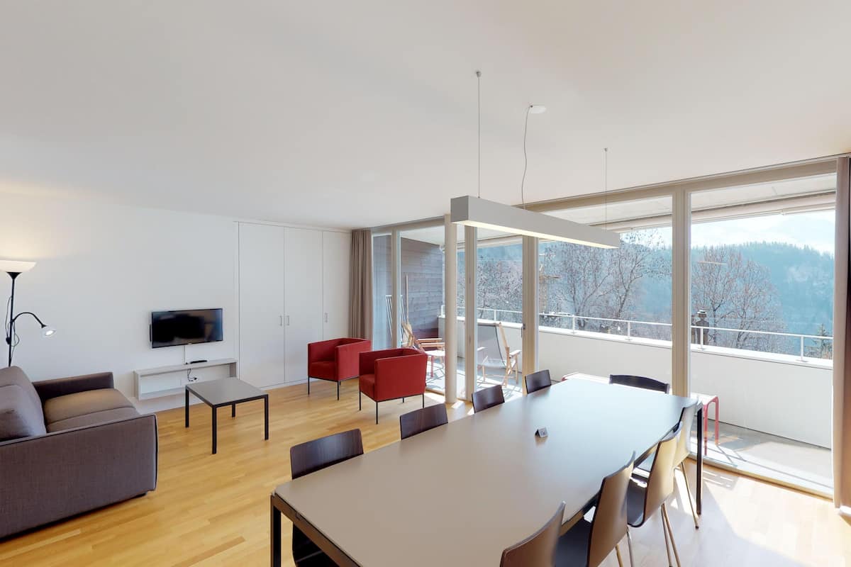 A modern living room with a large window, a flat-screen TV, a sofa, a dining table, and red chairs.