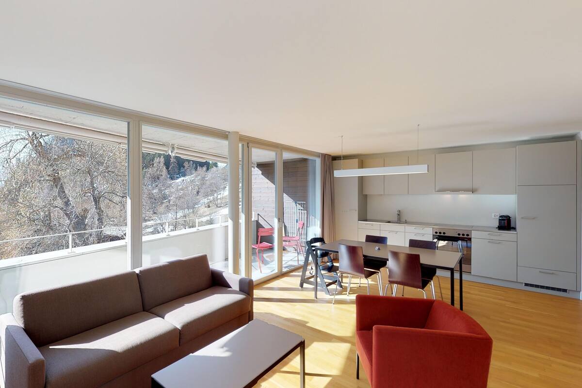 A modern living room with a sofa and chairs, a dining area with a table, and a kitchen with cabinets and a stove.