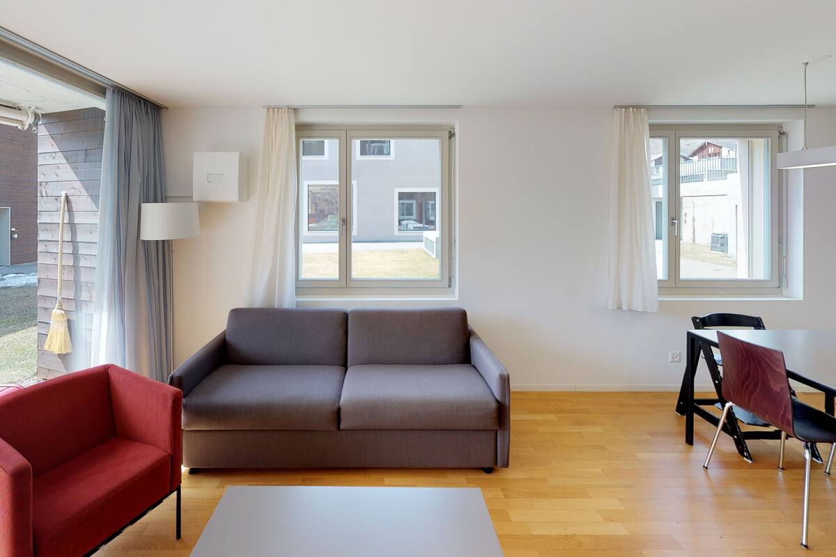 A modern living room with a grey sofa, a red armchair, a white coffee table, and a dining area with a table and chairs.