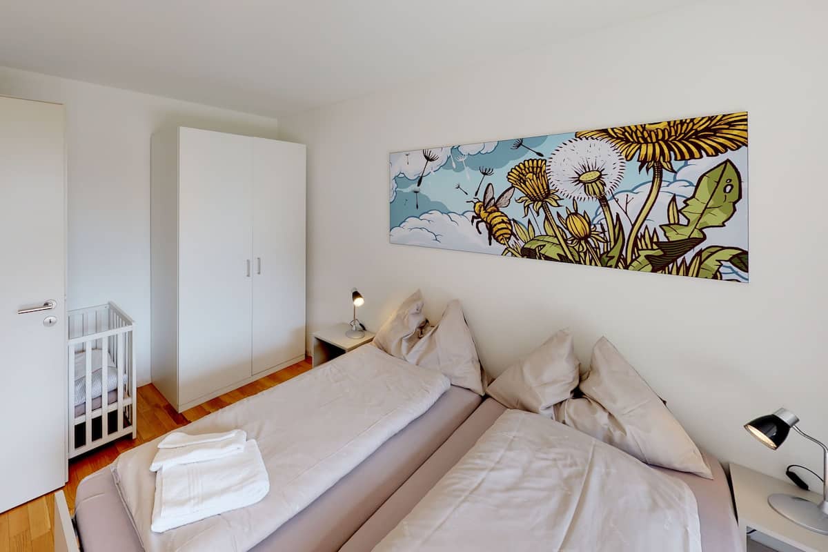 A bedroom with two beds, a crib, a nightstand with a lamp, and a wall painting.