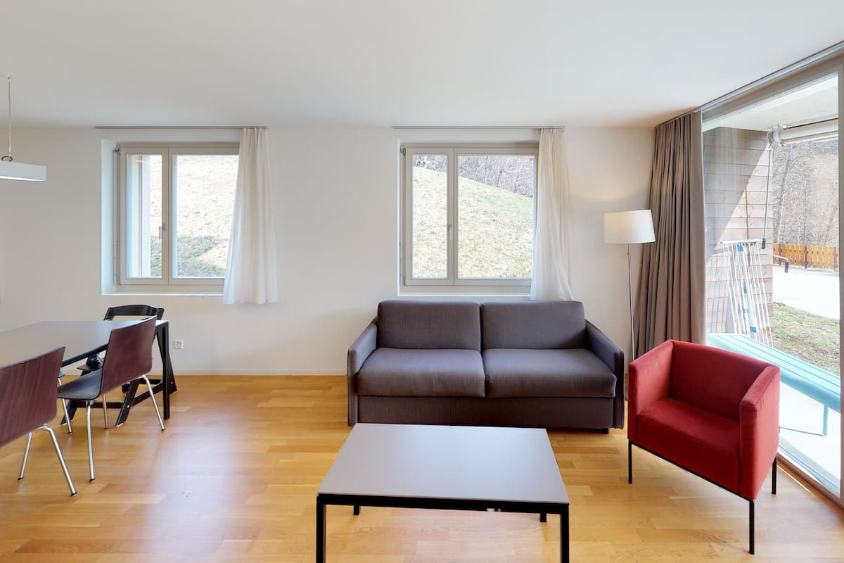 A modern living room with a sofa, a red armchair, a dining table with chairs, and large windows offering a view of the outdoors.