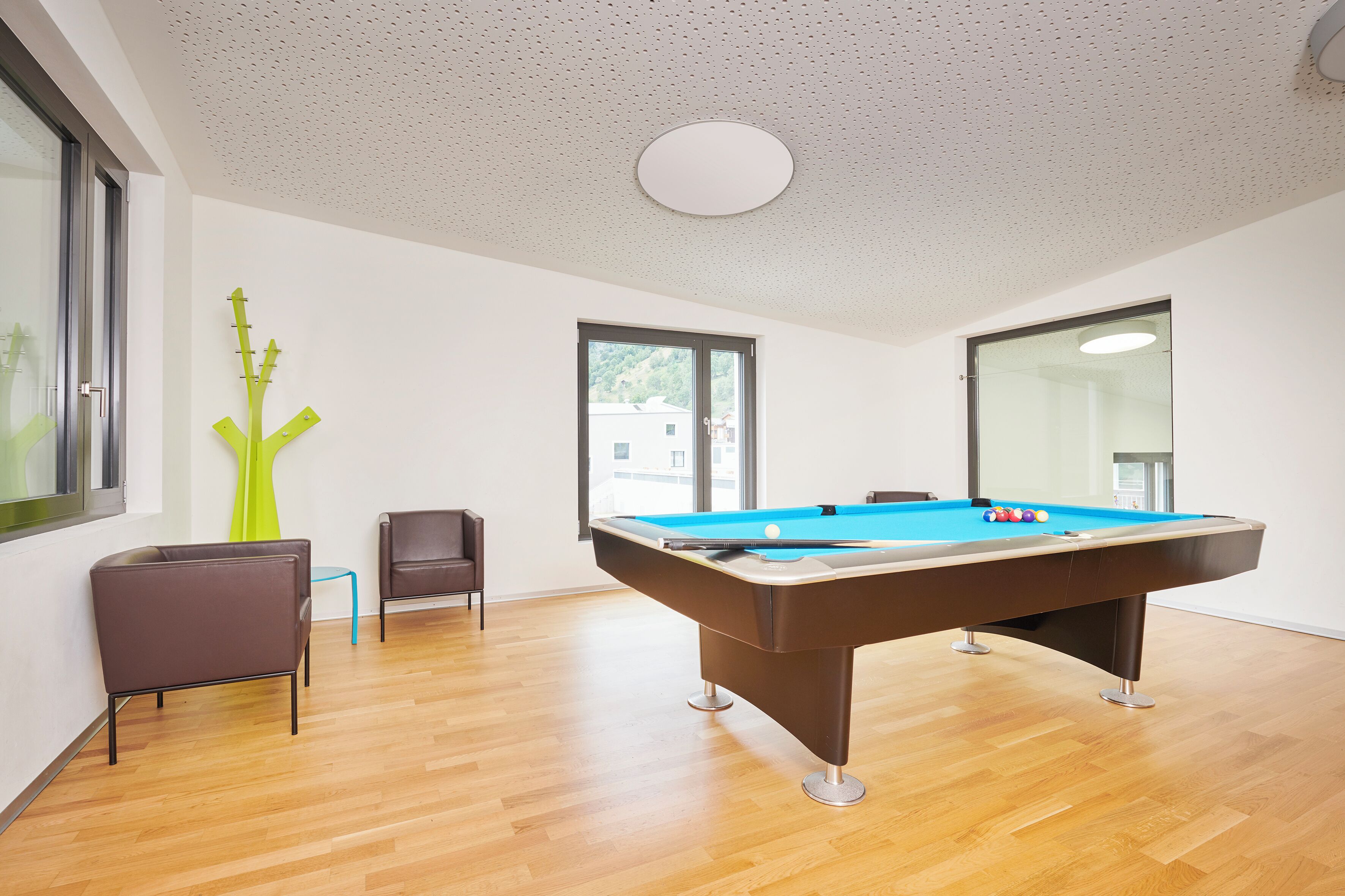 A modern room with a pool table, wooden flooring, a green abstract sculpture, and a window with a view of buildings.