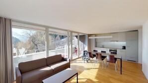 A modern living room with a sofa, a dining table with chairs, a kitchen area, and large windows offering a view of trees and mountains. - Reka-Feriendorf Blatten-Belalp (Naters)