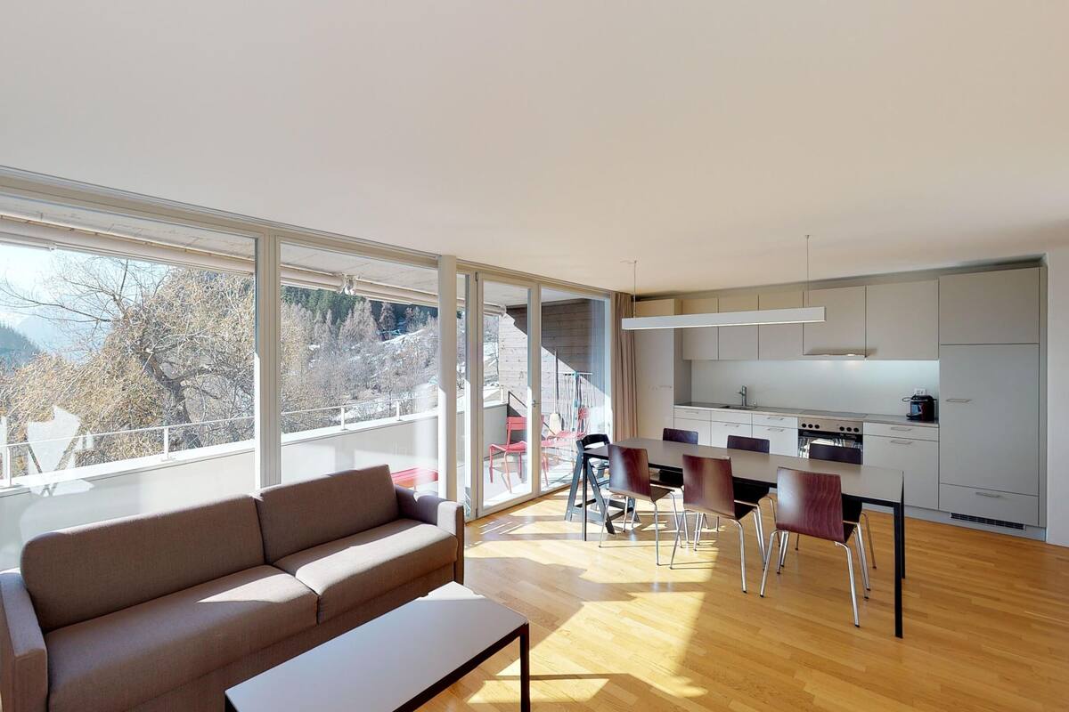 A modern living room with a sofa, a dining table with chairs, a kitchen area, and large windows offering a view of trees and mountains.