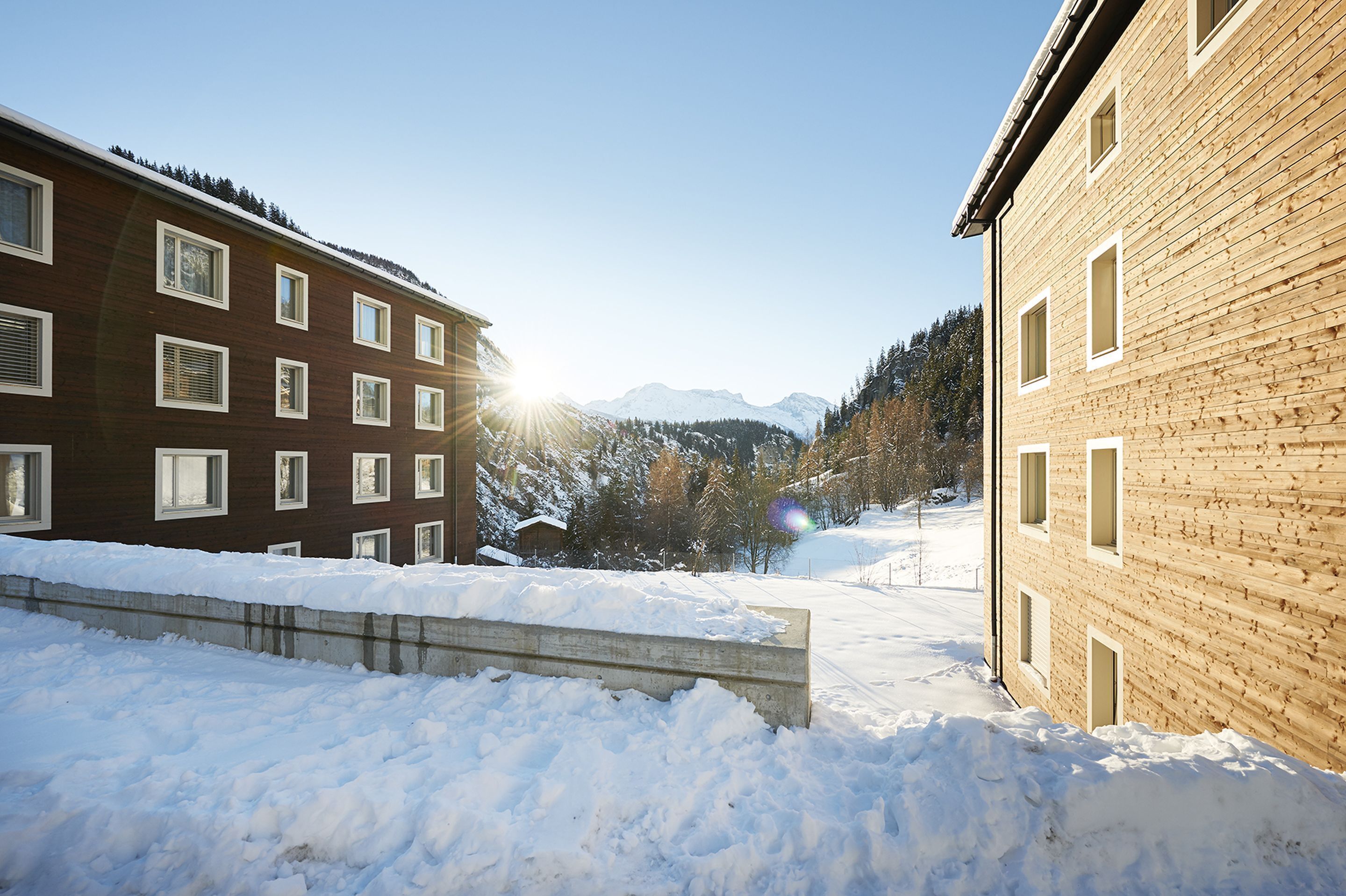 A snow-covered outdoor area with two buildings, one with a sloped roof and the other with a flat roof, both featuring multiple windows.