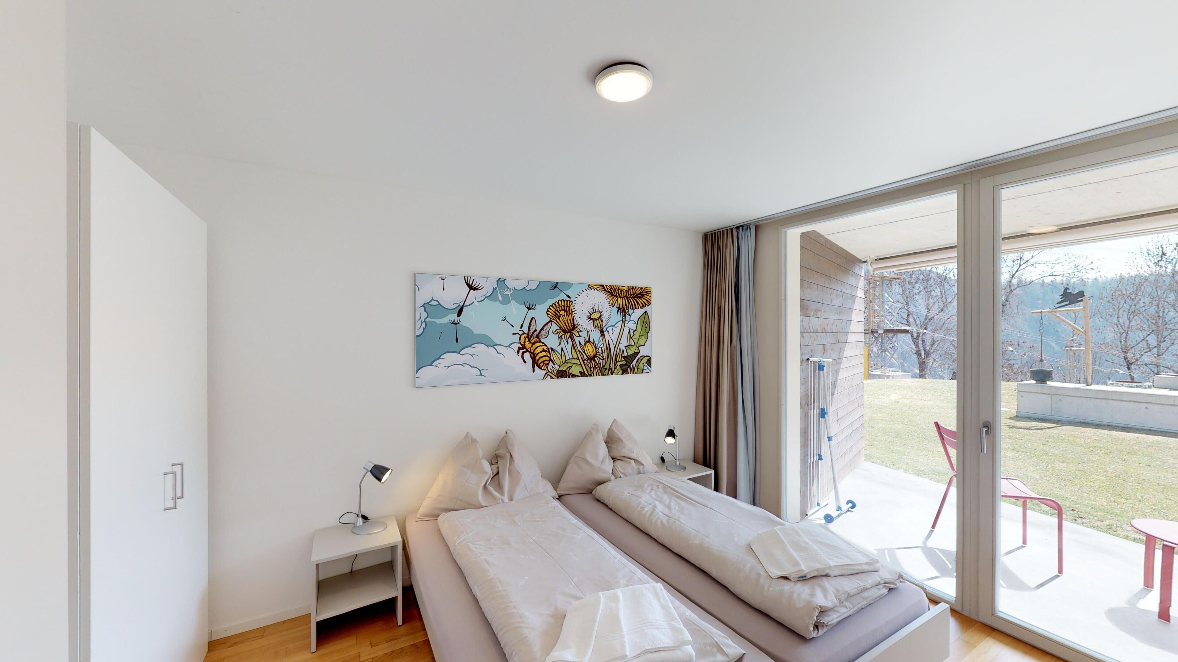 Une chambre moderne avec un grand lit, une table de chevet avec une lampe, une œuvre d’art accrochée au mur et une porte-fenêtre coulissante donnant sur un espace extérieur.