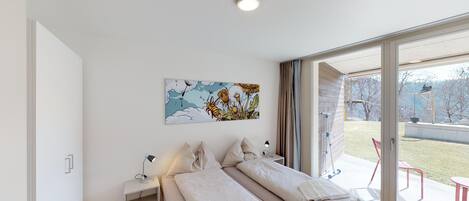 Une chambre moderne avec un grand lit, une table de chevet avec une lampe, une œuvre d’art accrochée au mur et une porte-fenêtre coulissante donnant sur un espace extérieur.
