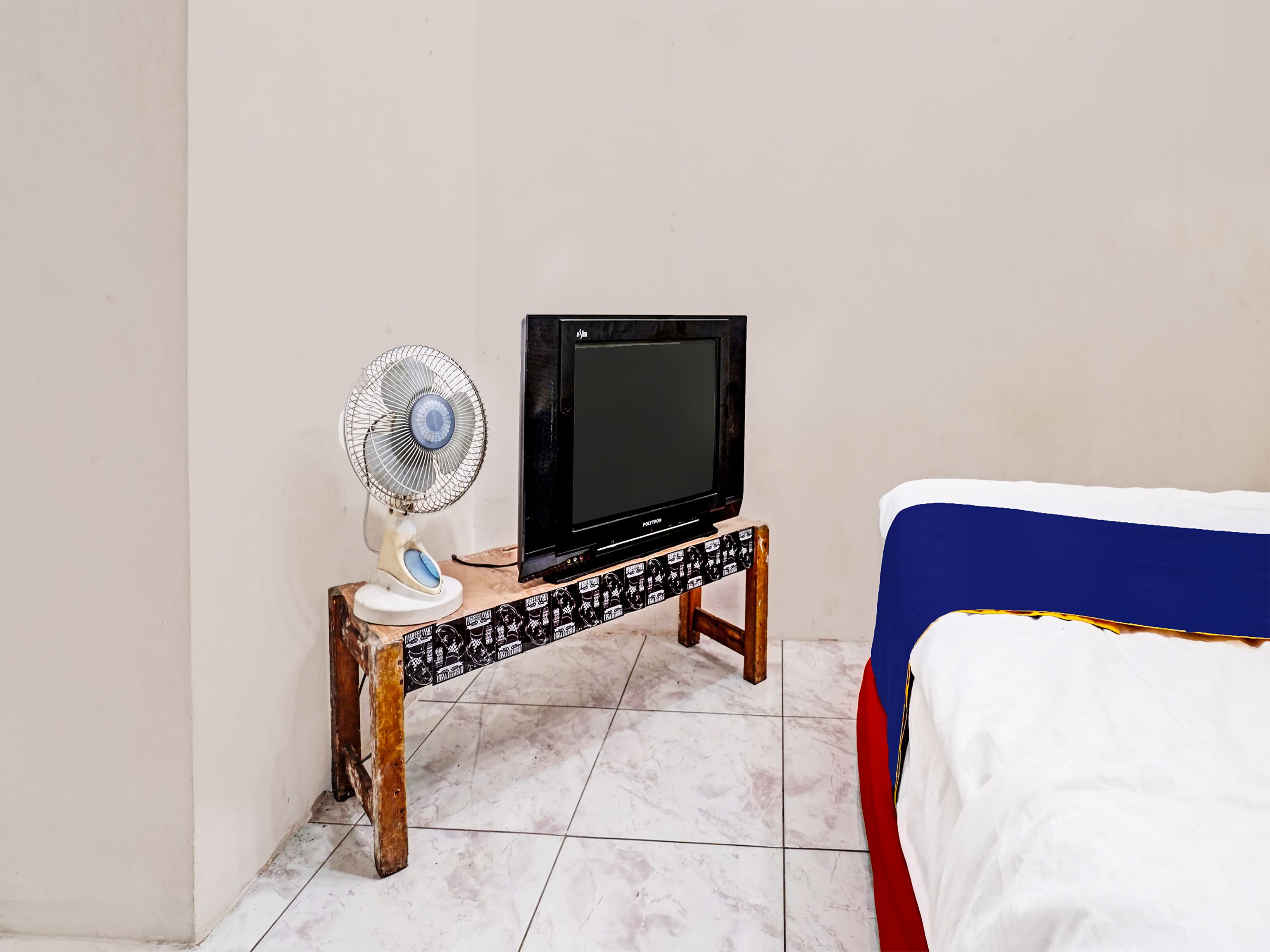Una habitación con una cama, un estante de madera que sostiene un televisor y un ventilador, y piso de baldosas.