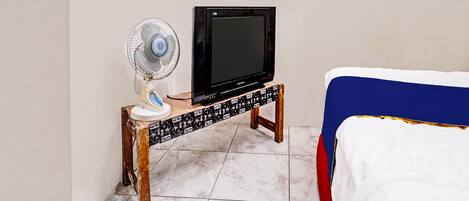 Una habitación con una cama, un estante de madera que sostiene un televisor y un ventilador, y piso de baldosas.