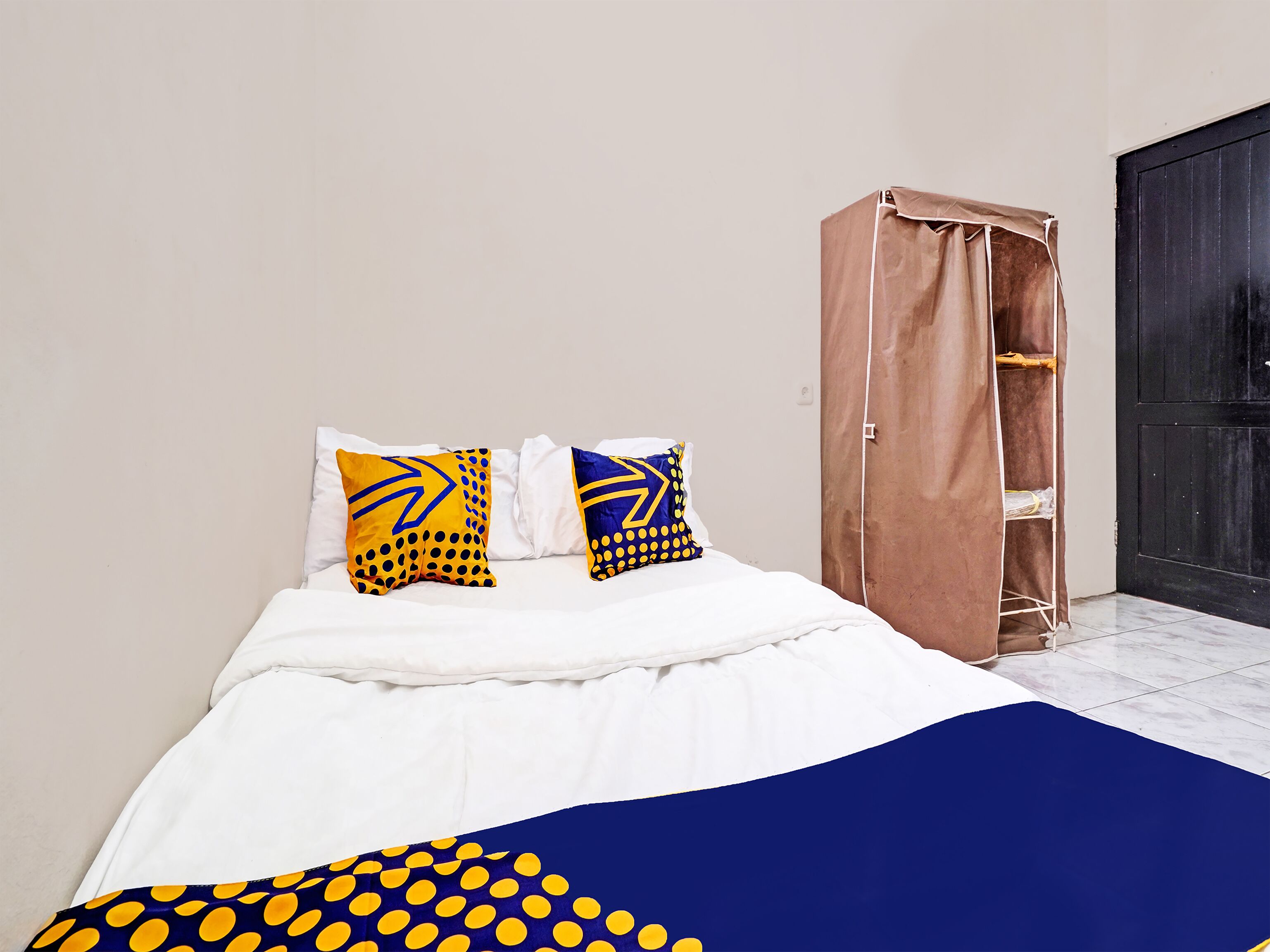 A bedroom with a bed, two pillows with yellow and blue patterns, a brown wardrobe, and a black door.