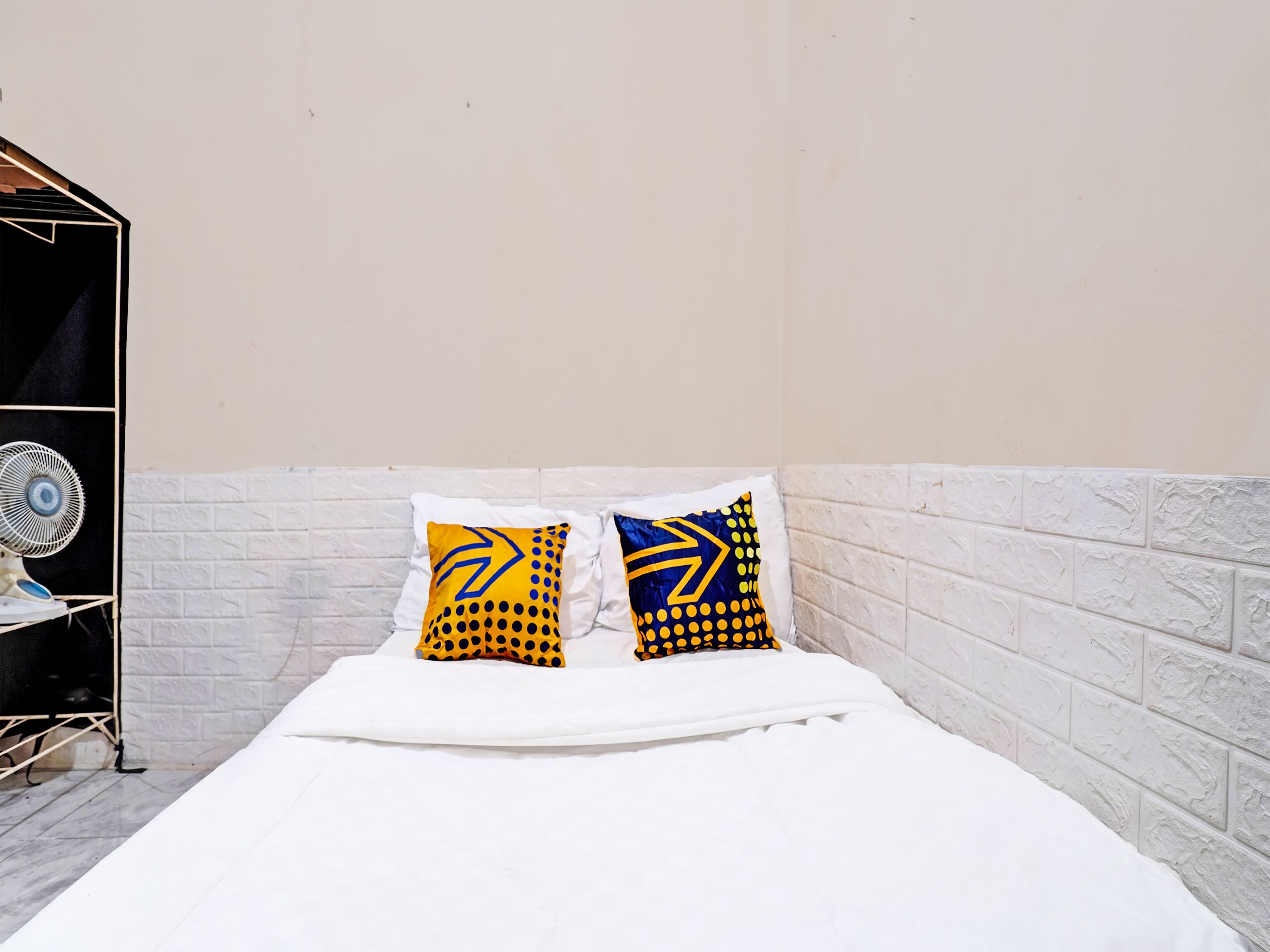 A bed with white bedding, two pillows with yellow and blue patterns, a white brick wall, and a white fan on a stand.