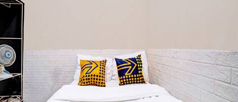 Una cama con ropa blanca, dos almohadas con diseños azules y amarillos, un muro de ladrillo blanco y un ventilador blanco sobre un soporte.