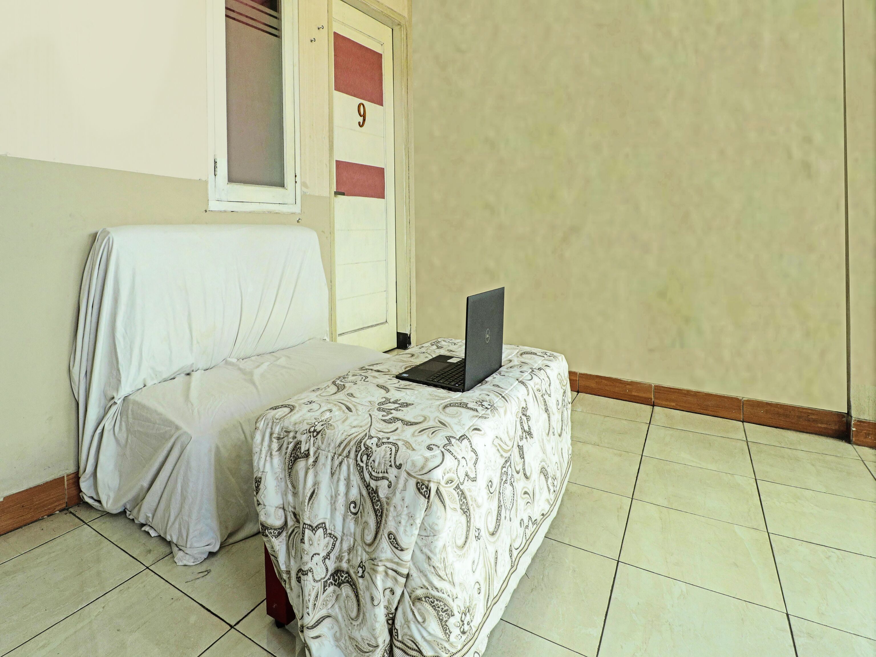 Ein Zimmer mit einem Bett, das mit einem weißen Laken bezogen ist, einem Laptop auf dem Bett, einer Tür mit der Nummer 9 und einem Fenster mit Blick ins Freie.