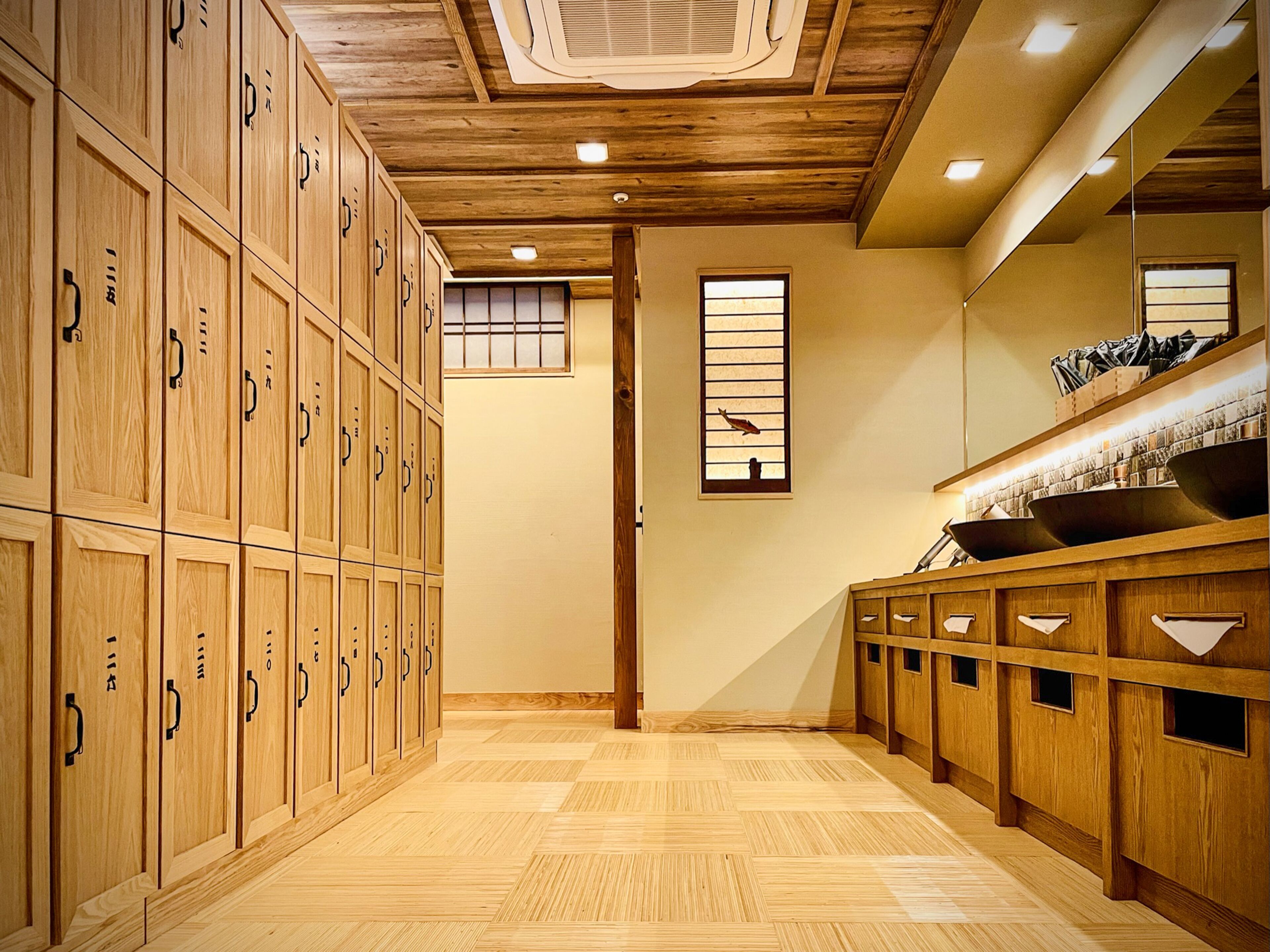 A hotel room with wooden lockers, a wooden floor, a window with a bird decoration, and a sink with a mirror above it.