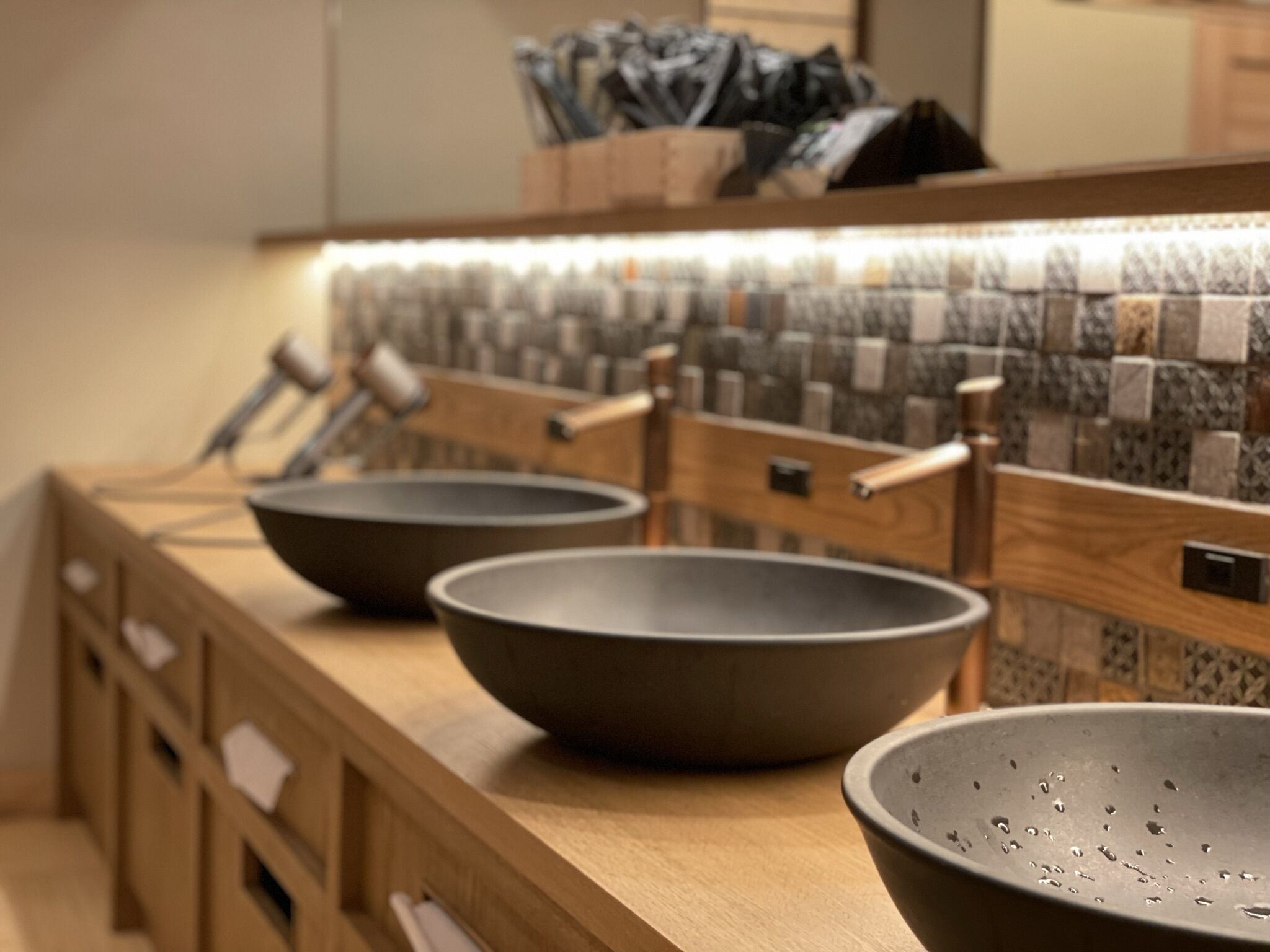 A kitchen with three dark bowls on a wooden countertop, a tiled backsplash, and wooden cabinets.
