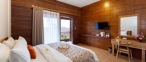 Habitación de hotel con pared de madera, cama con almohadas blancas y anaranjadas, escritorio con silla, televisor y ventana con cortinas.