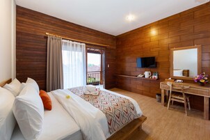 Habitación de hotel con pared de madera, cama con almohadas blancas y anaranjadas, escritorio con silla, televisor y ventana con cortinas.