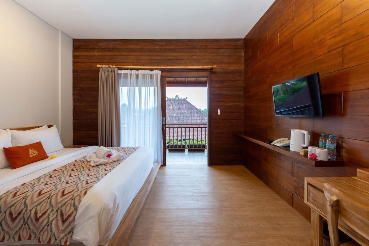Habitación de hotel con cama, televisor montado en la pared, piso de madera, balcón con vistas y una mesita con teléfono y artículos de aseo.