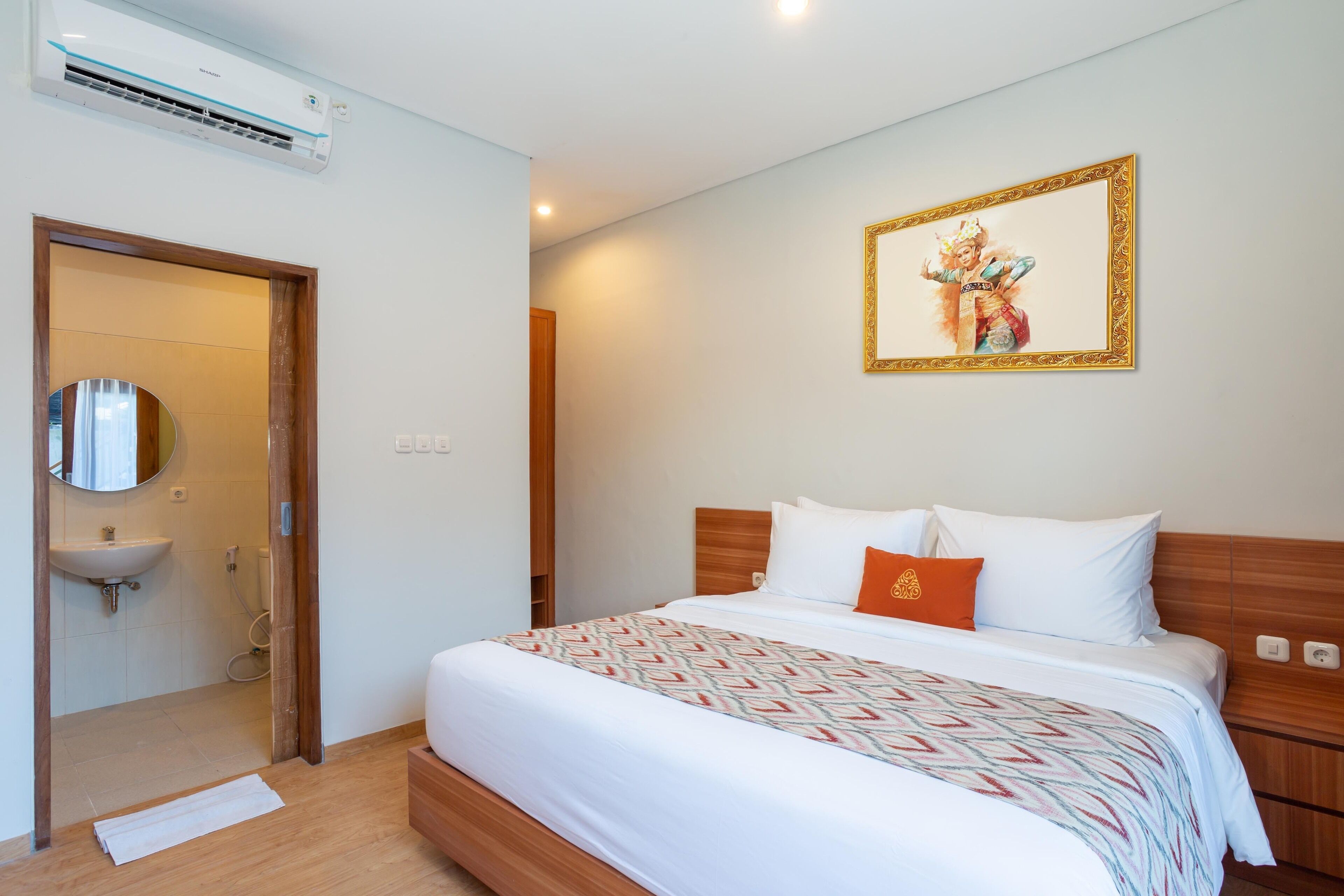 A hotel room with a bed, a wooden headboard, a wall-mounted air conditioner, a framed picture, and a bathroom with a sink and mirror.