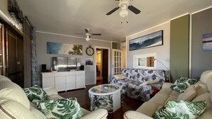 A living room with a sofa, coffee table, and ceiling fan. There is a kitchen area with cabinets and a refrigerator.