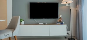 A modern living room with a flat-screen TV mounted on a white cabinet, a grey armchair, a small potted plant, a lamp, and a few books.