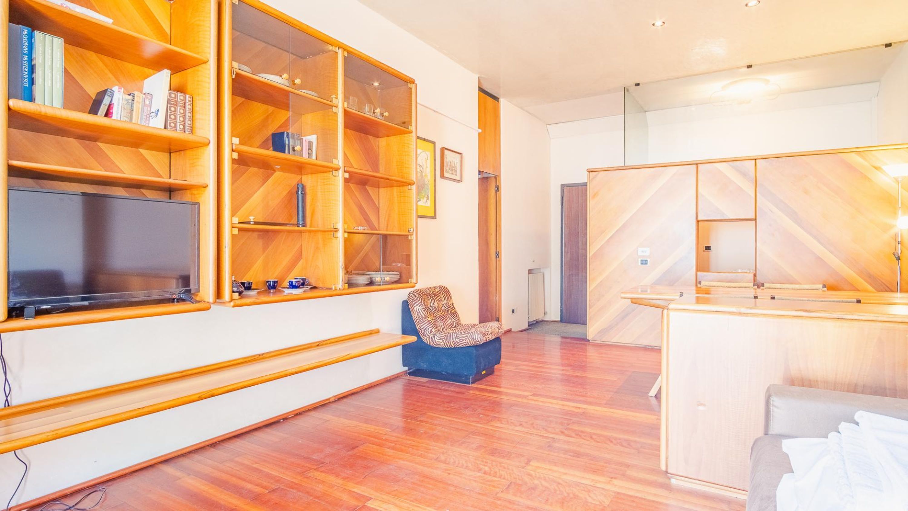 A modern living room with wooden shelving units, a flat-screen TV, a patterned armchair, and a wooden cabinet.