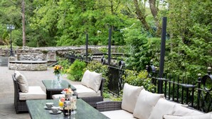 An outdoor seating area with wicker furniture and a glass table, surrounded by greenery and a stone wall.
