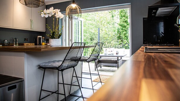 A modern kitchen with a wooden countertop, two black wire chairs, a hanging light fixture, and a view of greenery outside.