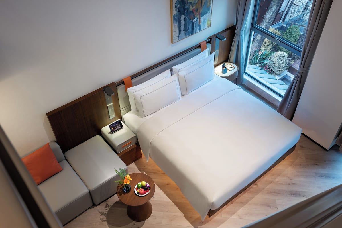 Une chambre d’hôtel moderne avec un grand lit, un petit canapé, une table de chevet sur laquelle se trouve un panier de fruits, et une fenêtre donnant sur un paysage verdoyant.