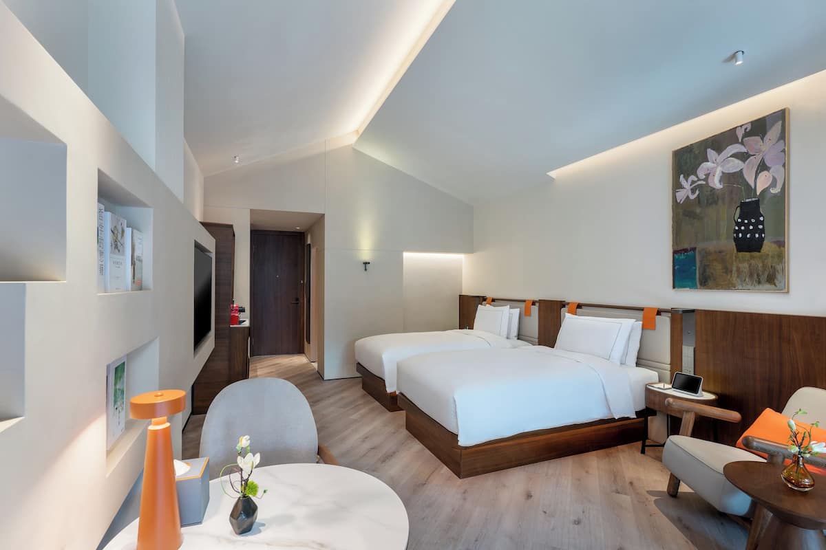 Une chambre d’hôtel avec deux lits, un bureau, une chaise, une petite table et un tableau au mur.