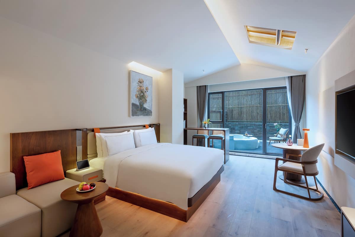 Une chambre d’hôtel moderne avec un grand lit, un canapé, une petite table avec des fruits, une chaise et une vue sur l’extérieur grâce à une grande fenêtre.