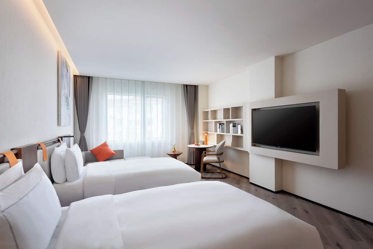 Une chambre d’hôtel avec deux lits, une télévision, une petite table avec une lampe et une bibliothèque.