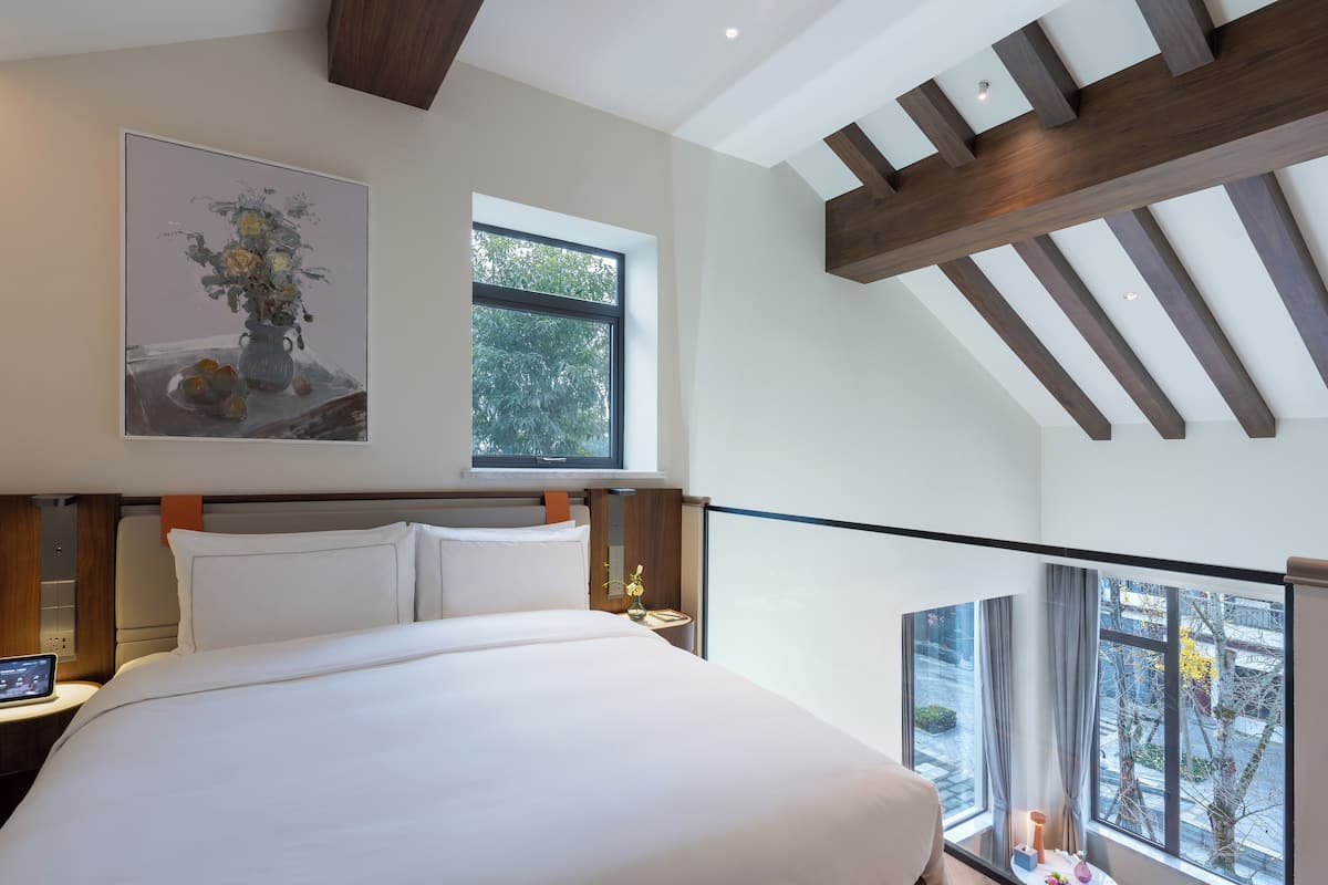 Une chambre d’hôtel moderne avec un grand lit, une table de chevet avec une lampe, un tableau au mur et une vue sur la ville grâce à de grandes fenêtres.