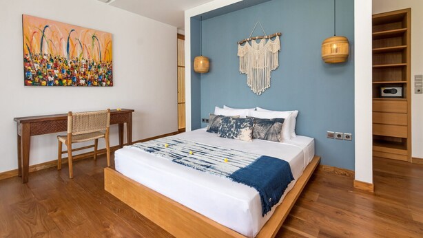 A bedroom with a wooden bed, a desk, a chair, a macrame wall hanging, and a wall painting. - Cheerful 4BR Villa W/ Pool & Balcony, Bali Villa 2219 (Kerobokan)